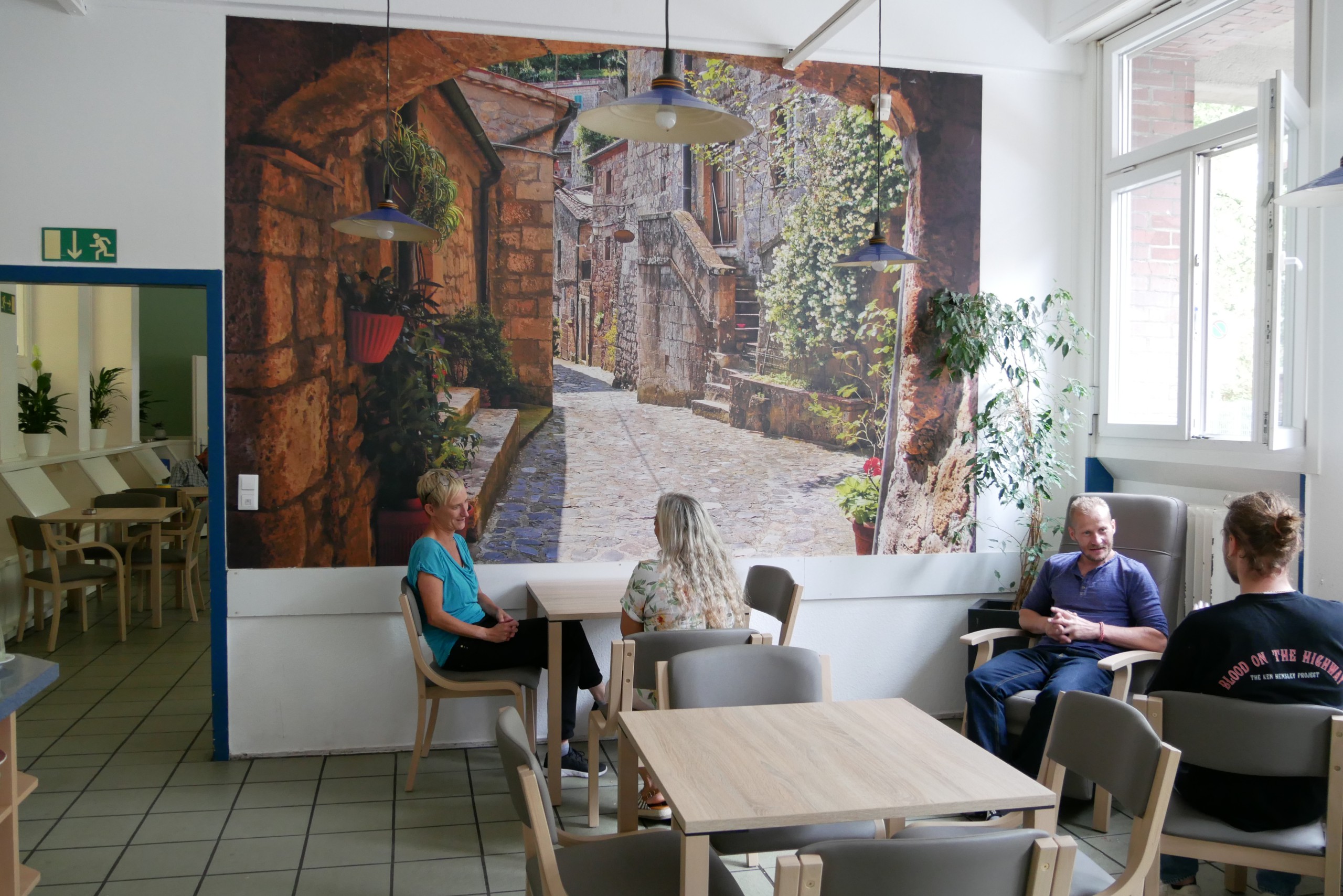 Menschen sitzen an kleinen Tischen in einem hellen Raum mit Fototapete an der Wand